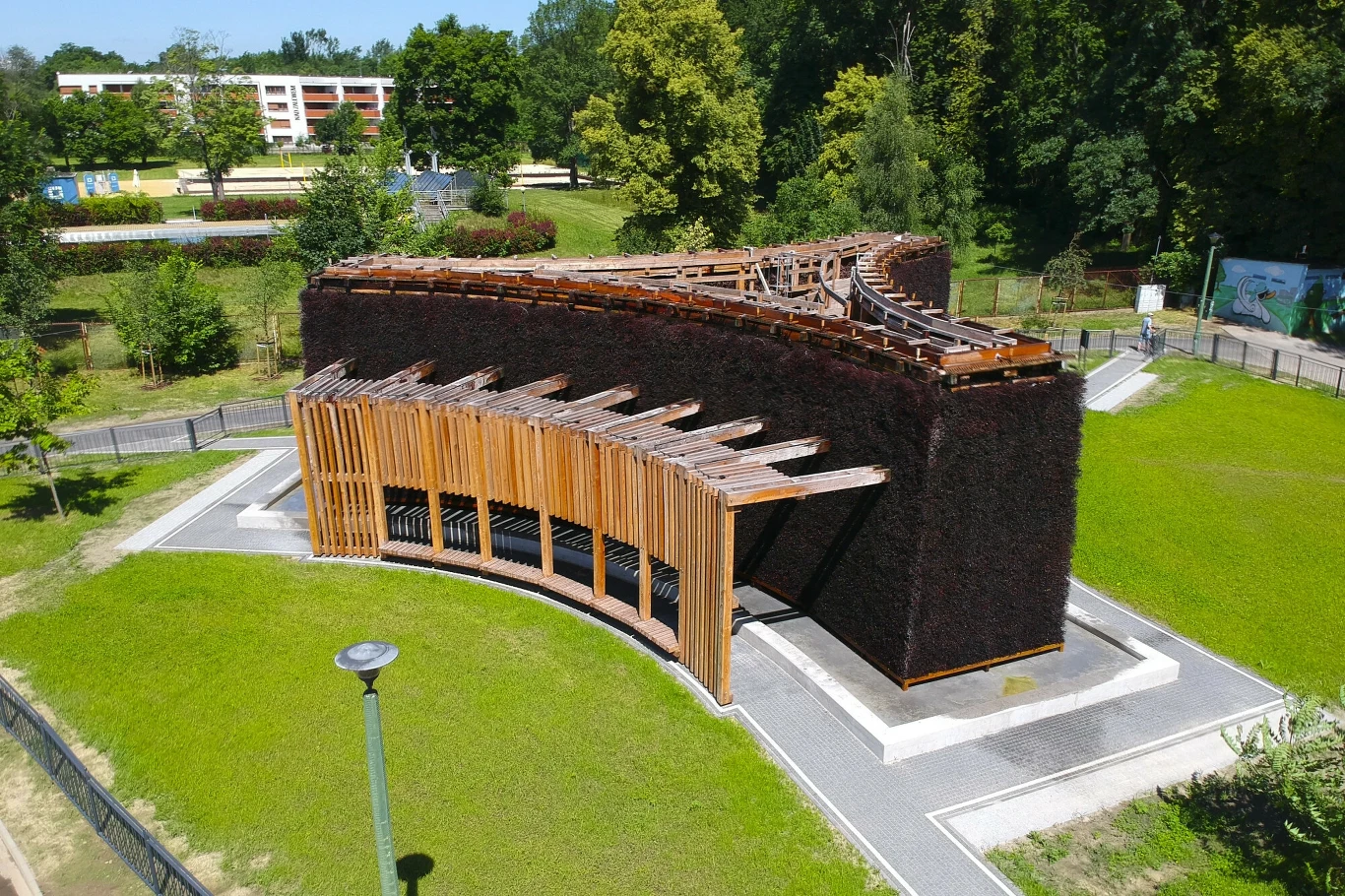 Tężnia przy Zalewie Nowohuckim pod okiem naukowców z Uniwersytetu Rolniczego w Krakowie Drewniana tężnia solankowa o nowoczesnej formie łuku otoczona trawnikiem i alejkami, w tle zabudowania oraz drzewa tworzące parkowy krajobraz.