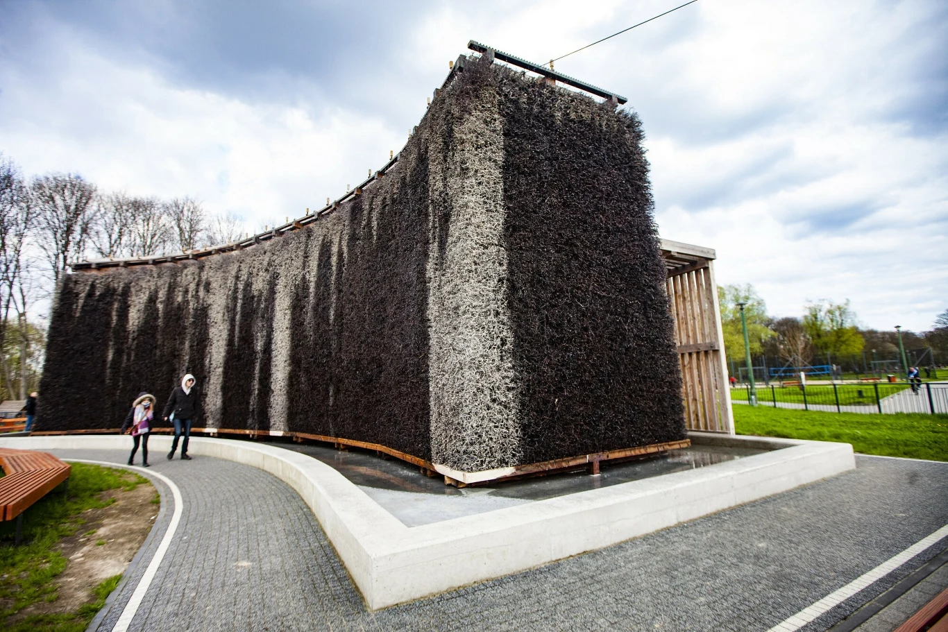 Stężenia bakterii i grzybów w solance tężni przy Zalewie Nowohuckim są niepokojące duża, zakrzywiona tężnia solankowa z ciemnymi gałęziami, otoczona alejkami spacerowymi, z dwoma osobami przechodzącymi obok oraz ławkami na trawniku, w tle drzewa i teren rekreacyjny