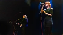 Dwie osoby wykonujące muzykę na scenie podczas koncertu, jedna kobieta śpiewa do mikrofonu, druga gra na instrumencie klawiszowym; wyraźne oświetlenie sceniczne podkreśla atmosferę występu.