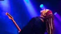 Muzyk z dredami, w opasce na głowie, grający na gitarze elektrycznej na scenie, w pełnym zaangażowaniu i silnym przechyleniu do tyłu, w tle widoczne bębny i intensywne światła sceniczne w odcieniach fioletu i niebieskiego.