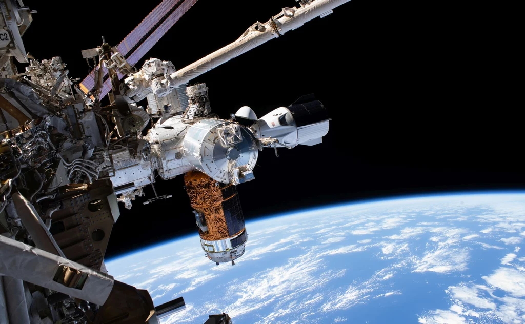 Kapsuła Dragon firmy SpaceX zadokowana do Międzynarodowej Stacji Kosmicznej. Stacja kosmiczna z przycumowanym statkiem kosmicznym na tle krzywizny Ziemi widocznej przez czarną przestrzeń kosmosu.