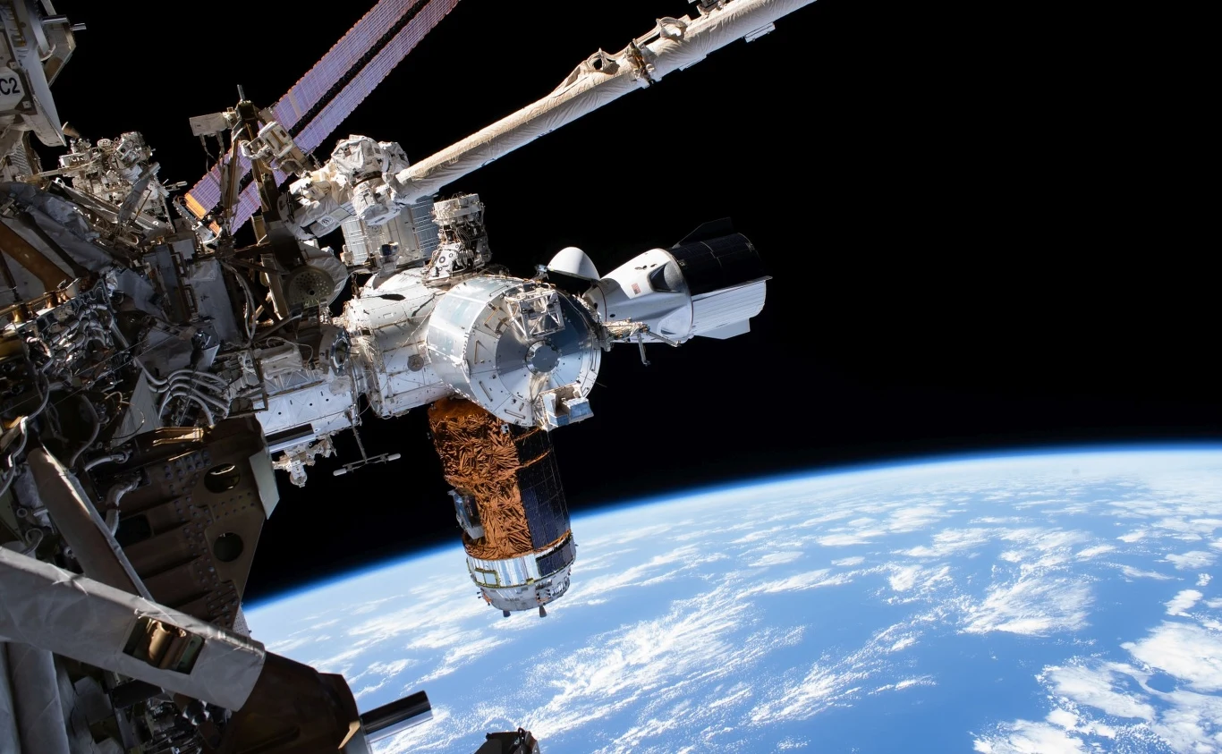 Stacja kosmiczna z przycumowanym statkiem kosmicznym na tle krzywizny Ziemi widocznej przez czarną przestrzeń kosmosu.