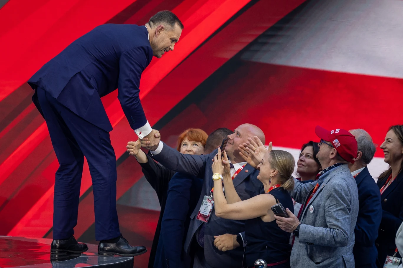 Przeciwko Nawrockiemu stosuje się prymitywne i stereotypowe chwyty Mężczyzna ubrany w ciemny garnitur stoi na podwyższeniu, pochyla się i ściska ręce kilku osób zgromadzonych poniżej, z entuzjazmem wyciągających dłonie w jego stronę; tło utrzymane jest w czerwonych i białych barwach.