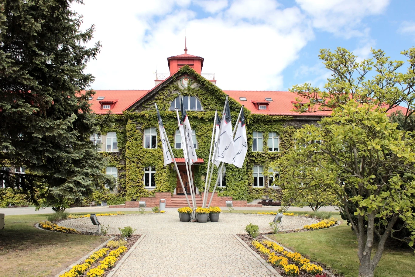 Historyczny budynek Fabryki Samochodów Ciężarowych STAR w Starachowicach Historyczny budynek z czerwonym dachem porośnięty zielonym bluszczem, przed wejściem sześć flag ustawionych w donicach, przed budynkiem brukowana alejka otoczona żółtymi kwiatami i zielonymi drzewami, niebo częściowo zachmurzone.