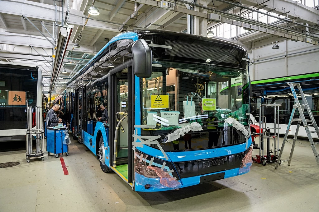 Nowy MAN w fabryce w Starachowicach Nowoczesny, niebieski autobus znajduje się wewnątrz hali produkcyjnej podczas montażu, przy pojeździe pracuje kilku techników, a wokół widoczne są drabiny, narzędzia oraz elementy wyposażenia fabryki.