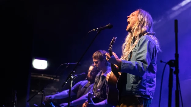 Trzech mężczyzn występuje na scenie podczas koncertu, wokalista z długimi blond włosami śpiewa do mikrofonu i gra na gitarze akustycznej, w tle widoczne instrumenty klawiszowe oraz niebieskie światła sceniczne.