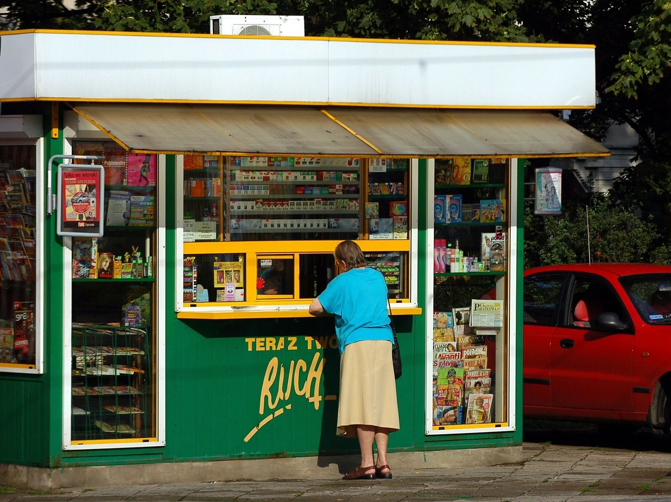 Starsza kobieta ubrana w niebieską bluzkę i beżową spódnicę stoi przy okienku zielonego kiosku z prasą i artykułami spożywczymi, obok zaparkowany jest czerwony samochód.