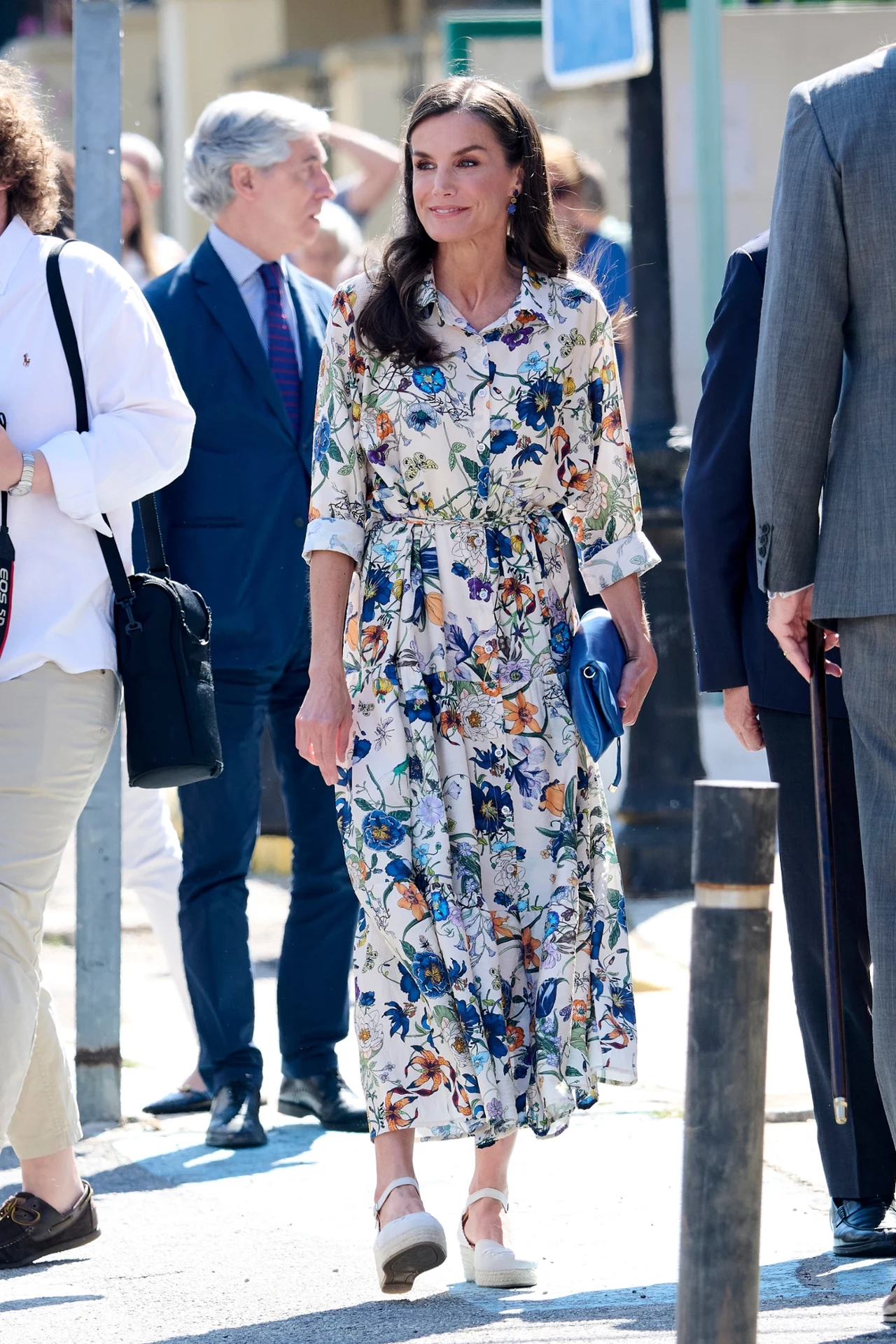Elegancka kobieta w długiej sukience w kolorowe kwiaty, trzymająca niebieską kopertówkę, spaceruje po ulicy w towarzystwie kilku osób w ubraniach formalnych.