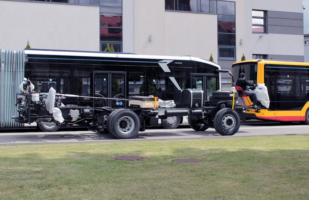 W fabryce autobusów MAN w Starachowicach produkowane są też gotowe podwozia pod zabudowę Szkielet autobusu na podwoziu w trakcie montażu, ustawiony na tle nowoczesnego autobusu przegubowego i żółto-czerwonego autobusu miejskiego, oba zaparkowane przy ulicy na tle budynku mieszkalnego.
