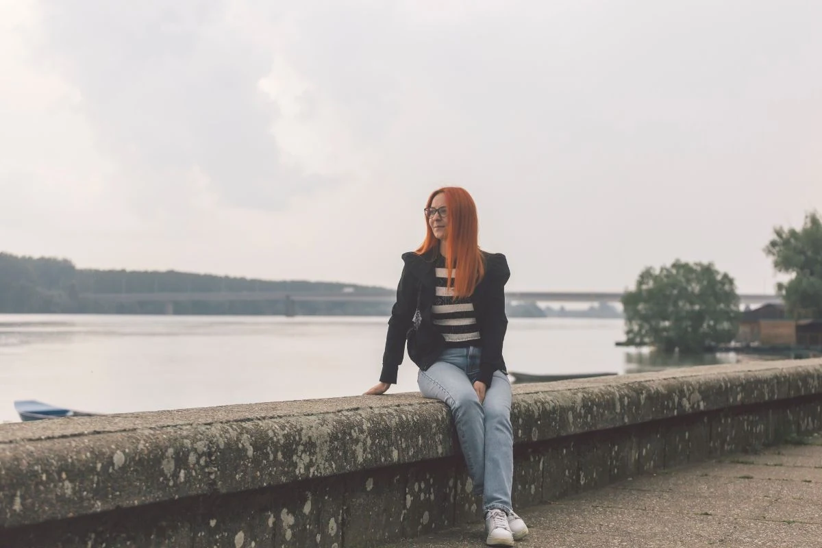 Kobieta z rudymi włosami ubrana w czarną marynarkę, w paskowaną bluzkę i dżinsy, siedzi na murku nad brzegiem rzeki, w tle widać drzewa, most oraz łódkę na wodzie.
