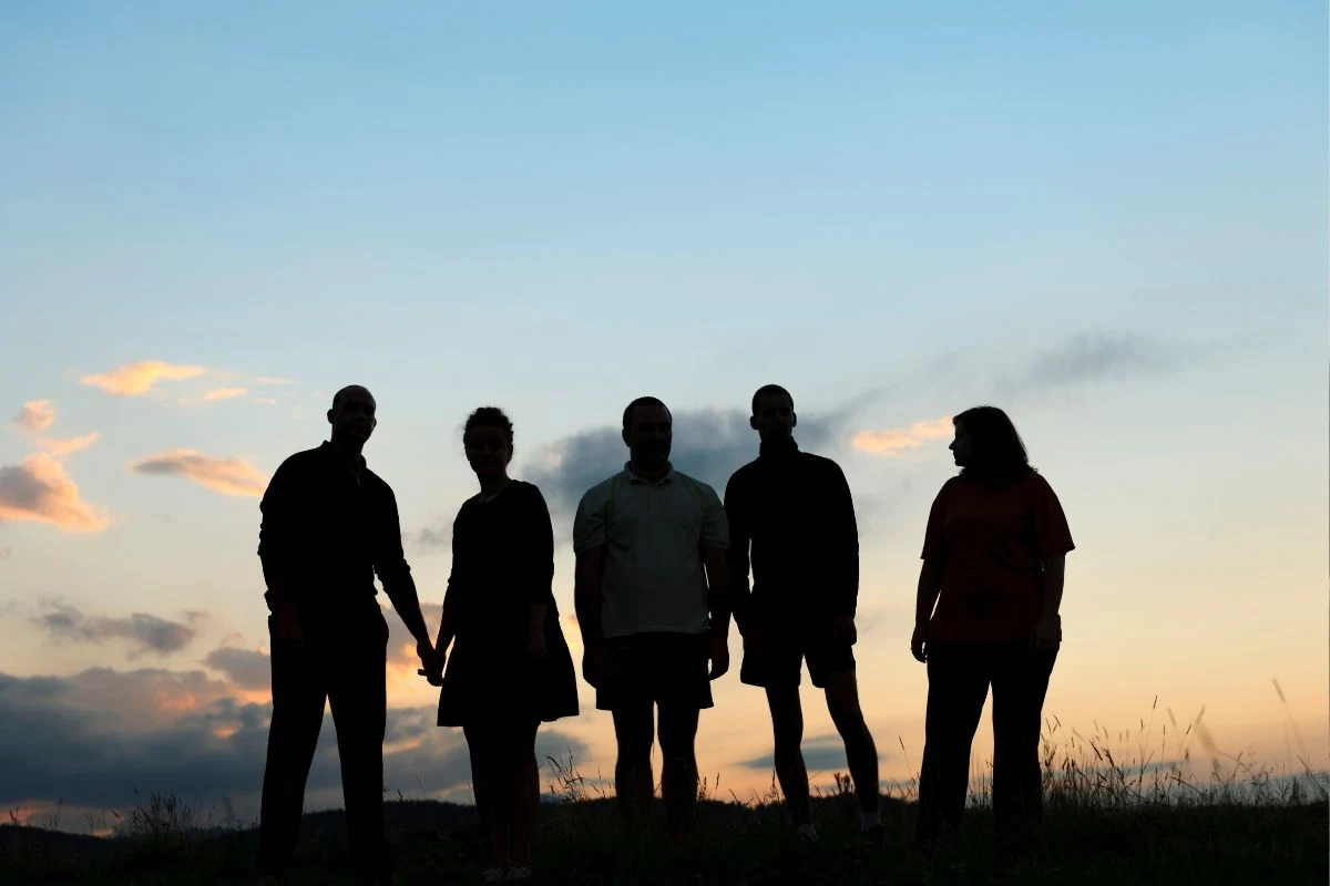 Pięć osób stoi obok siebie na tle zachodzącego słońca, sylwetki ludzi są ciemne i wyraźnie odcinają się od jasnego, kolorowego nieba, na twarzach brak rozpoznawalnych szczegółów.