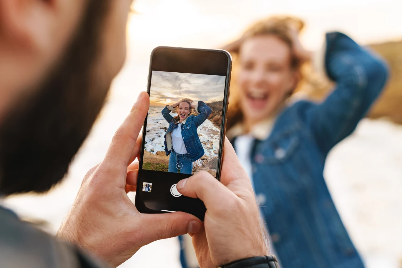 Mobilne studio fotograficzne w twojej kieszeni. Najlepsze aplikacje do edycji zdjęć Osoba uśmiechnięta w kurtce jeansowej pozuje do zdjęcia na świeżym powietrzu, drugi człowiek fotografuje ją smartfonem z bliskiej odległości, w tle widoczny krajobraz nad wodą.