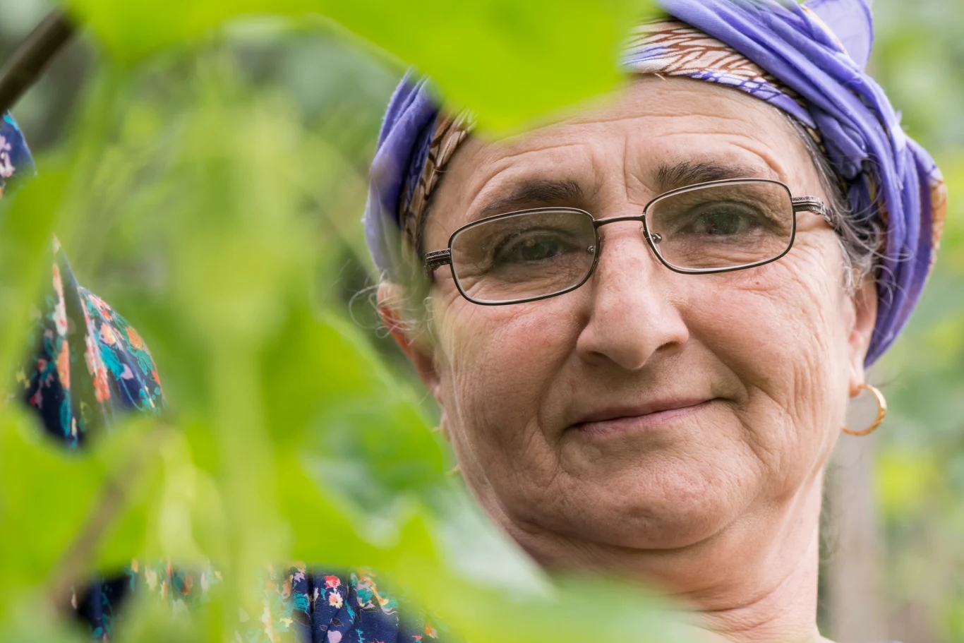 Horoskop finansowy od wróżki Anne. Sprawdź, co nas czeka Ujęcie starszej kobiety w okularach oraz chuście na głowie, spoglądającej łagodnie do obiektywu, w otoczeniu zielonych liści.