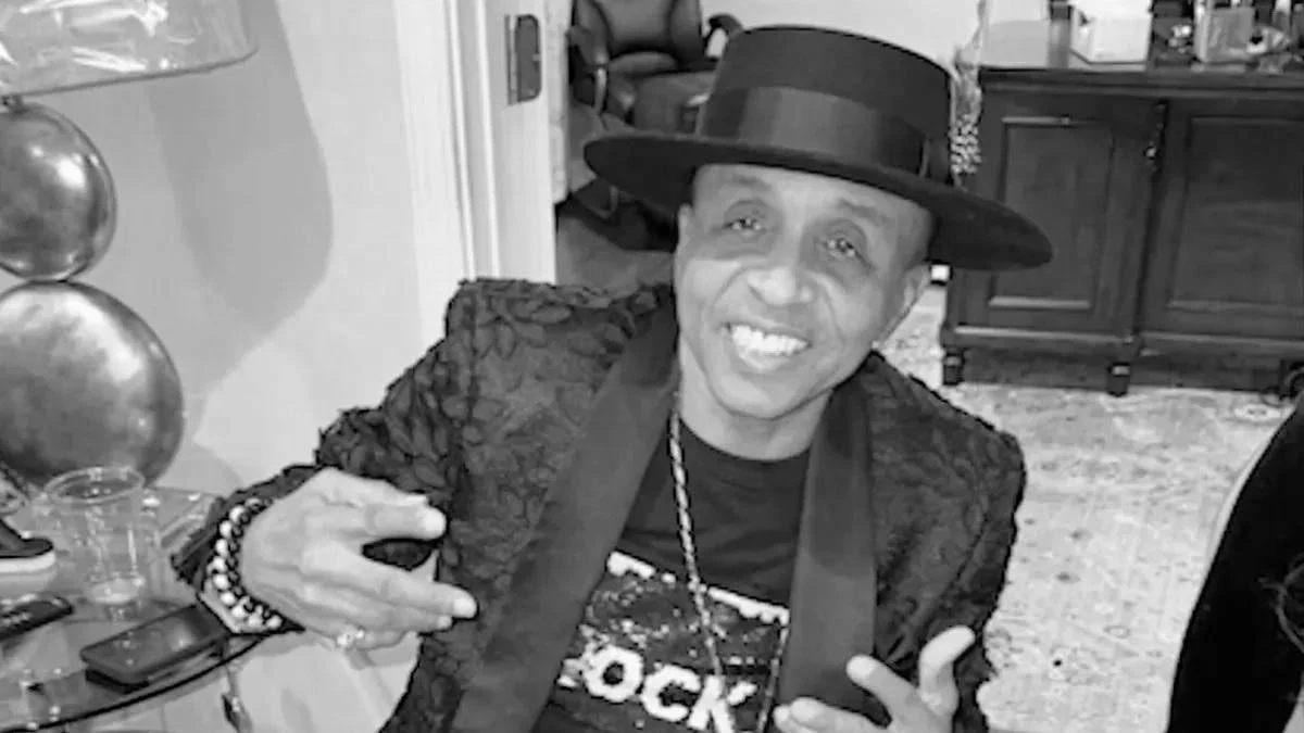 Michael Sumler (Kool & The Gang) miał 71 lat Uśmiechnięty mężczyzna w kapeluszu i eleganckiej marynarce, siedzący przy stole, pokazuje gest dłoni, w tle widoczne są meble i szklanki.