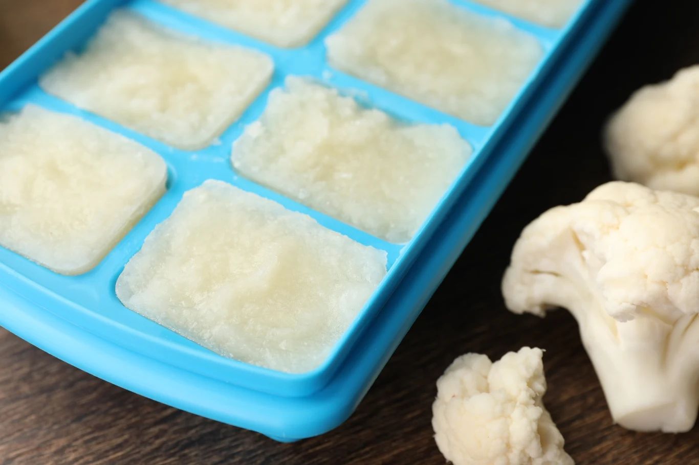 Foremka do lodu z zamrożonym puree kalafiorowym na drewnianym blacie, obok kawałki świeżego kalafiora.