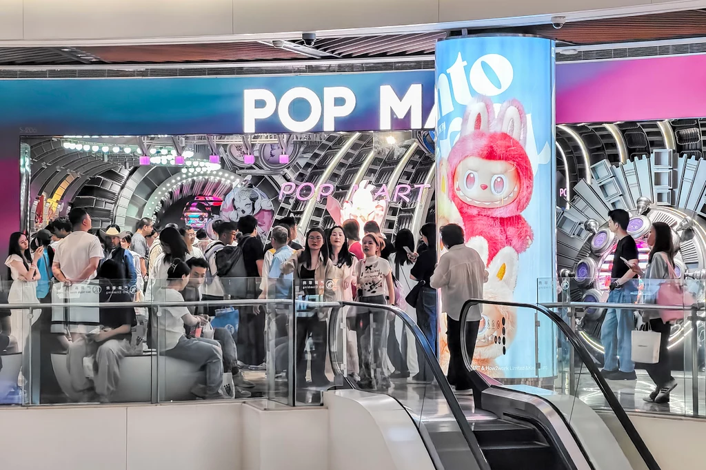Pod sklepami Pop Mart tworzą się wielogodzinne kolejki, by zakupić choć jedną lalkę Labubu Tłum ludzi gromadzący się przed kolorowo oświetlonym wejściem do sklepu lub miejsca rozrywki z nazwą POP MART, jasne neony, nowoczesne wnętrze, duży różowy pluszowy królik na podświetlanej kolumnie.