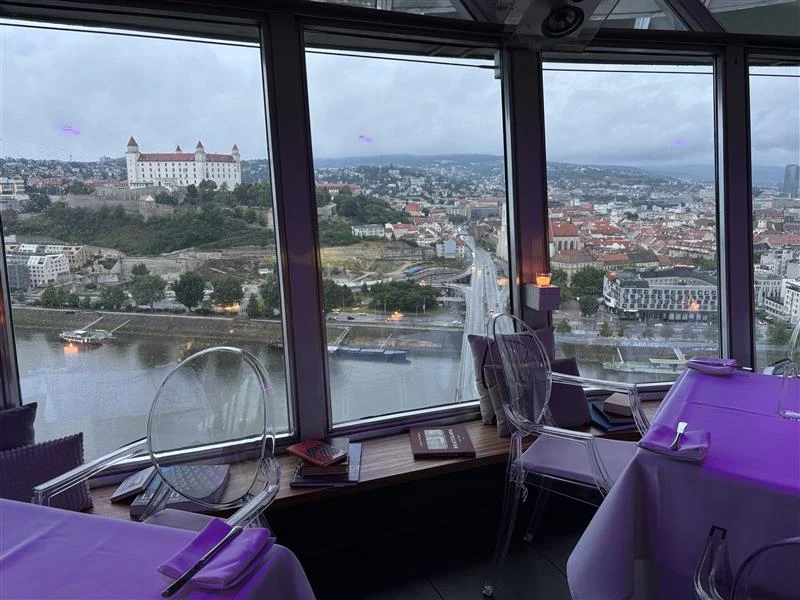 Widok z restauracji UFO Widok z panoramicznego okna restauracji na miasto z zamkiem na wzgórzu po lewej stronie i rzeką oraz mostem biegnącym przez środek kadru; nowoczesne wnętrze z nakrytymi stolikami i przezroczystymi krzesłami.
