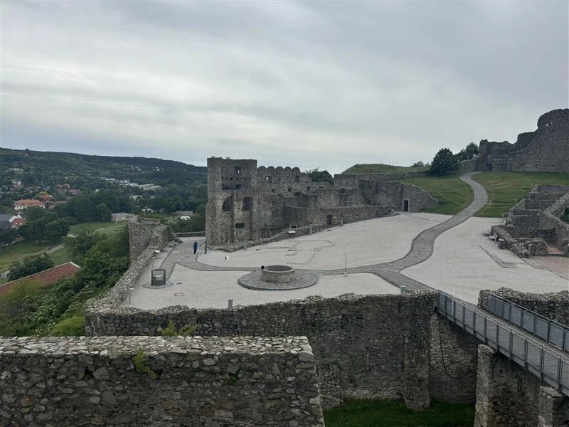 Zamek Devin Ruiny średniowiecznego zamku z kamiennymi murami, dziedziniec z zachowanymi fragmentami fortyfikacji, ścieżką prowadzącą w górę oraz rozległym widokiem na zielone wzgórza i miasteczko w tle.