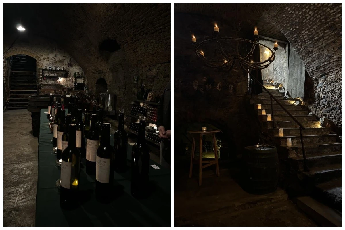Winnice Svätý Jur Stół z butelkami wina ustawiony w zabytkowej, kamiennej piwnicy, obok widoczne stare schody prowadzące na wyższy poziom, oświetlenie klimatyczne, surowe mury z cegły, na ścianie wisząca stylowa żyrandolowa lampa.