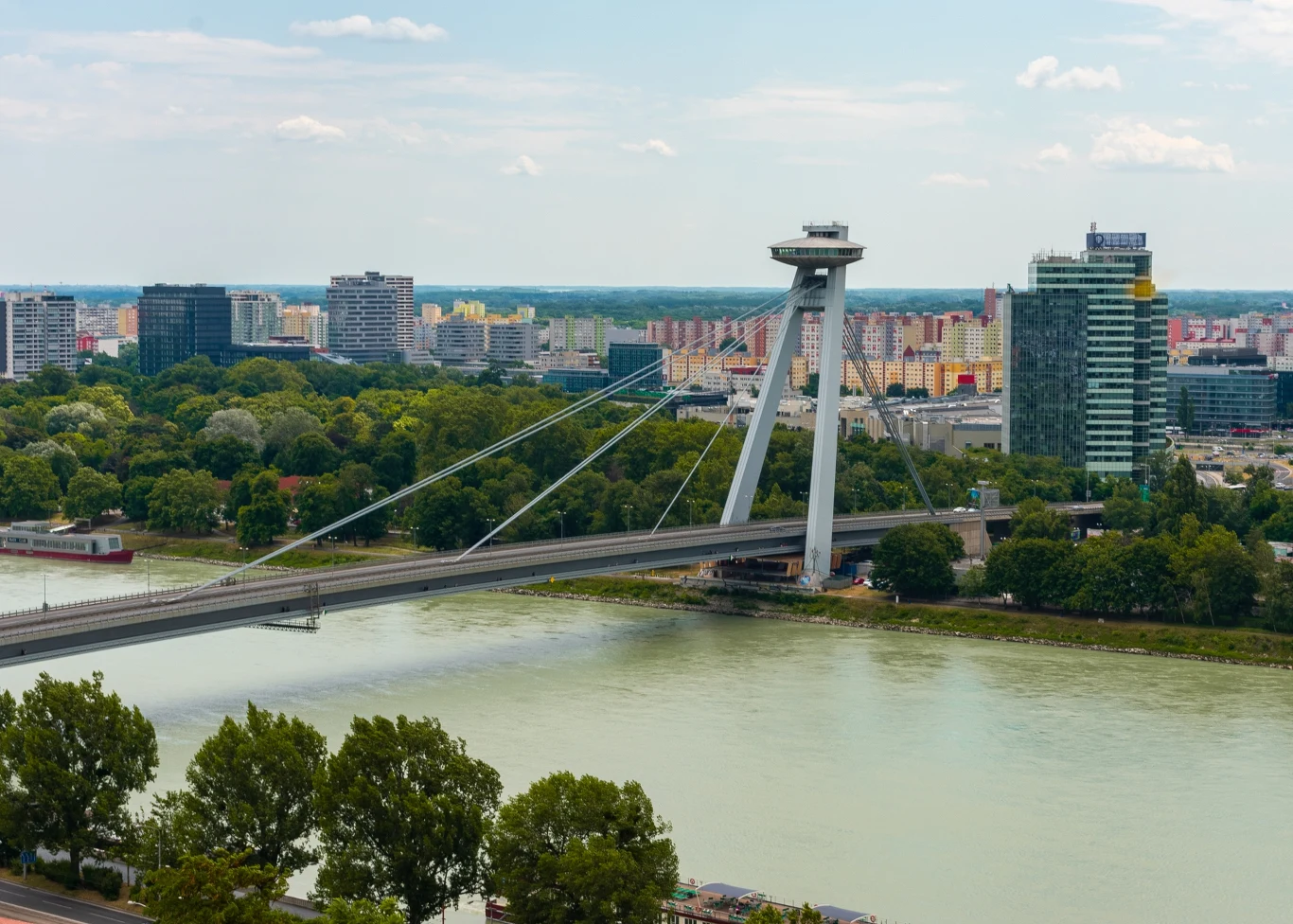 UFO Bratysława Most wantowy nad rzeką z charakterystyczną wieżą przypominającą talerz, w tle nowoczesne budynki i wysokie apartamentowce, dookoła widoczna zieleń drzew i zabudowa miejska.