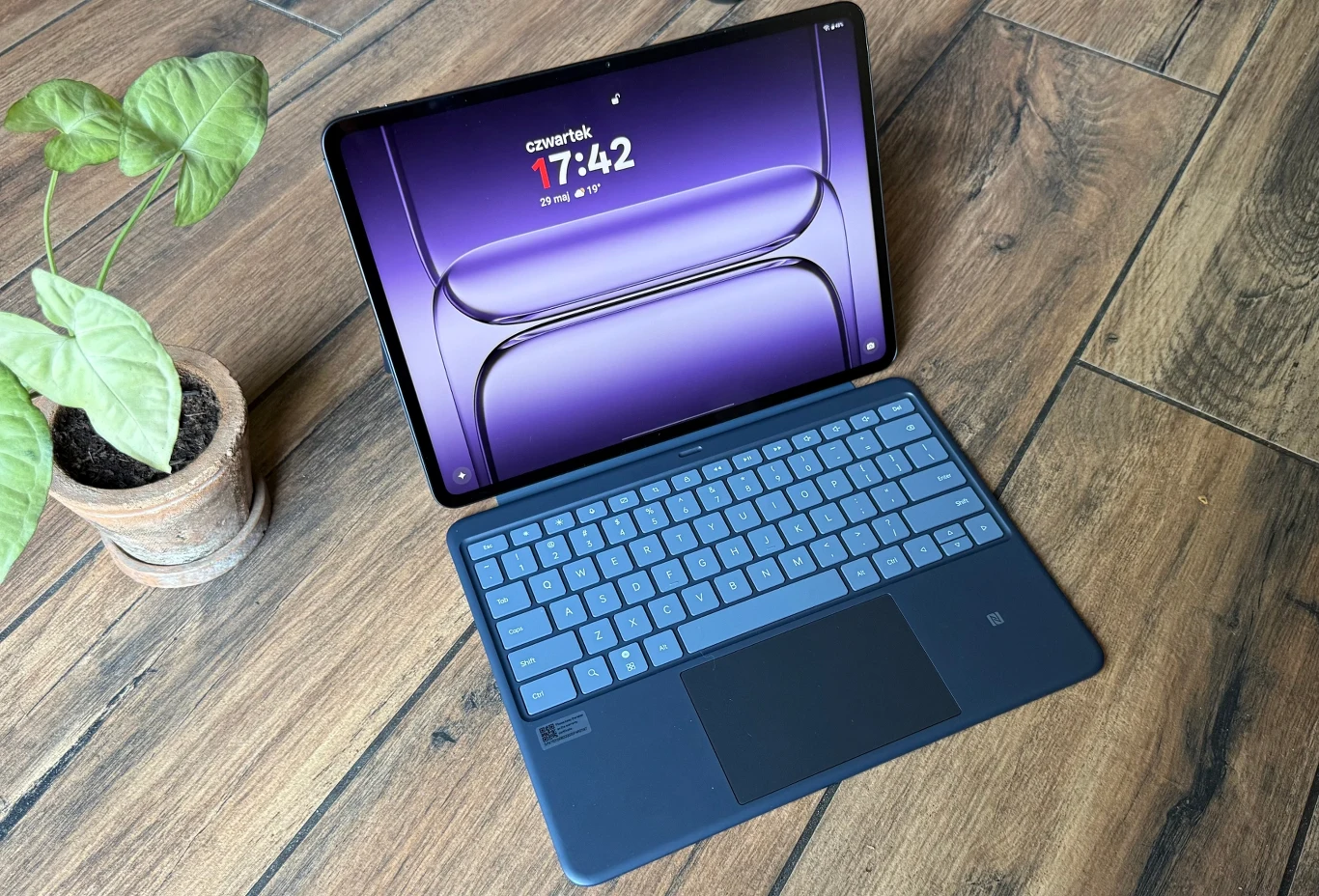Test OnePlus Pad 3. Czy to najlepszy tablet z Androidem? Tablet z podłączoną klawiaturą ustawiony na drewnianej podłodze obok doniczki z zieloną rośliną, wyświetlający ekran główny z godziną.