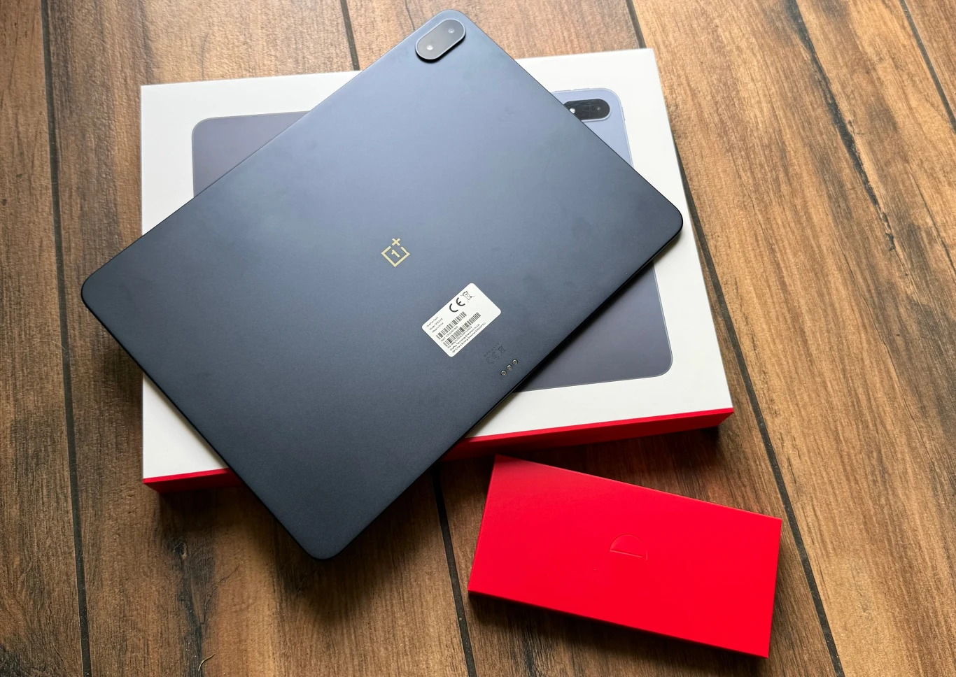 Zawartość opakowania z tabletem OnePlus Pad 3. Tablet OnePlus w kolorze czarnym leżący na białym opakowaniu z czerwonymi akcentami, obok znajduje się czerwone pudełko, całość umieszczona na drewnianej podłodze. Na tablecie widoczna naklejka producenta i aparat w rogu.