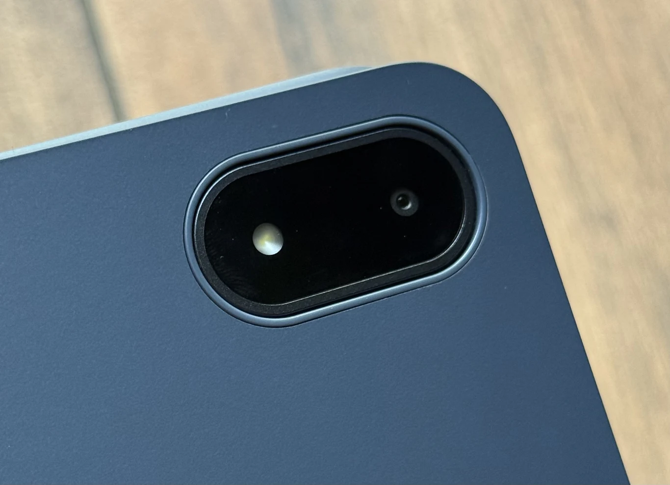 Aparat tabletu OnePlus Pad 3 nie jest mocną stroną urządzenia. Zbliżenie na tylną kamerę tabletu w metalowej obudowie, z widoczną soczewką aparatu i lampą błyskową na tle rozmytego drewnianego podłoża.