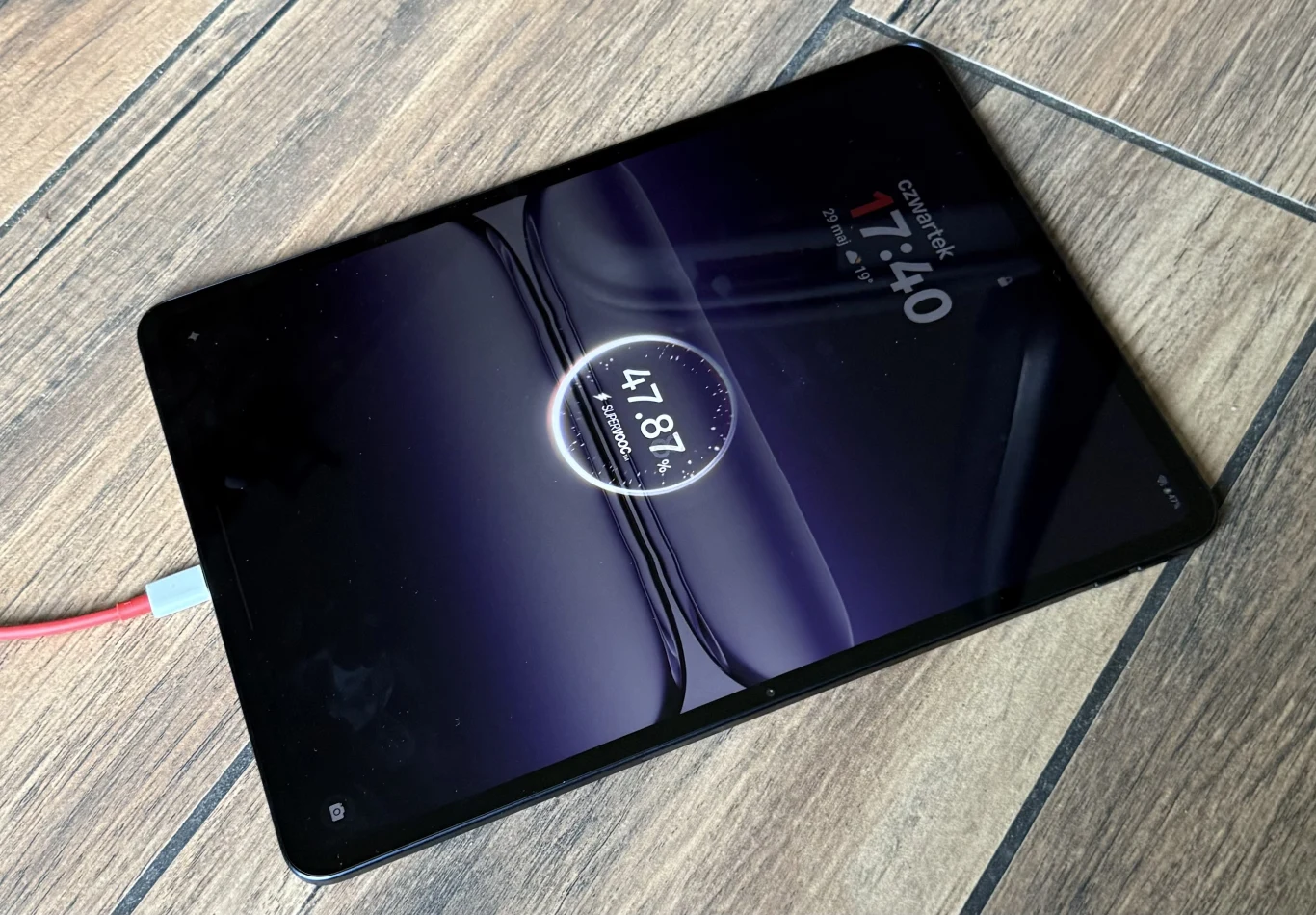 OnePlus Pad 3 wspiera szybkie ładowanie SuperVOOC o mocy 80 W. Tablet ładowany kablem USB-C leży na drewnianej podłodze, na ekranie wyświetlany jest stan baterii oraz aktualna godzina.