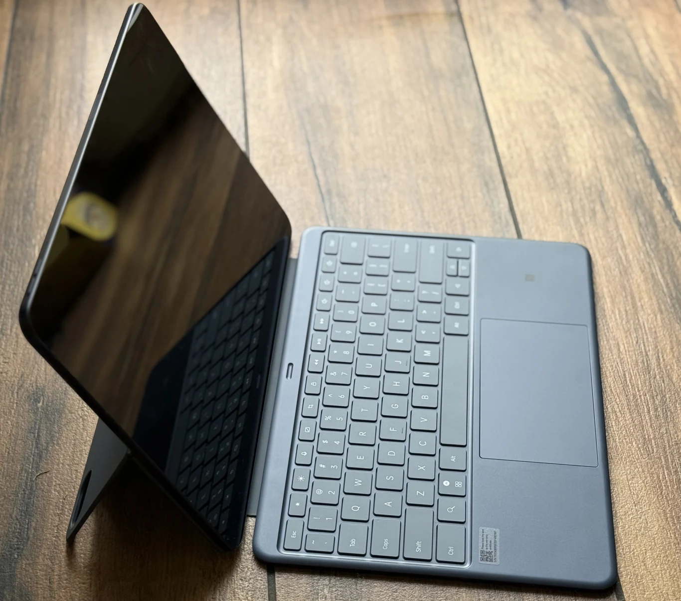 Dzięki dedykowanym akcesoriom OnePlus Pad 3 jest w stanie zastąpić laptopa. Tablet z podłączoną klawiaturą na stojaku, ustawiony na drewnianej powierzchni, ekran tabletu jest wygaszony lub ciemny.