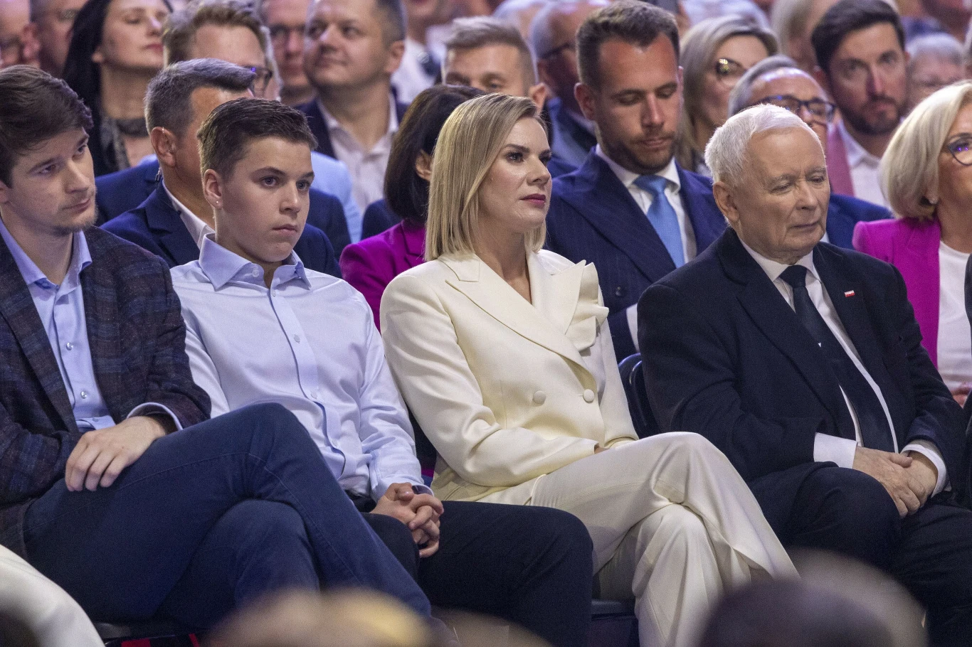 Marta Nawrocka w Łodzi Grupa dorosłych osób i młodzieży siedzi w pierwszym rzędzie na dużym spotkaniu, skupione i elegancko ubrane, w tle widoczne kolejne rzędy obserwujących ludzi.