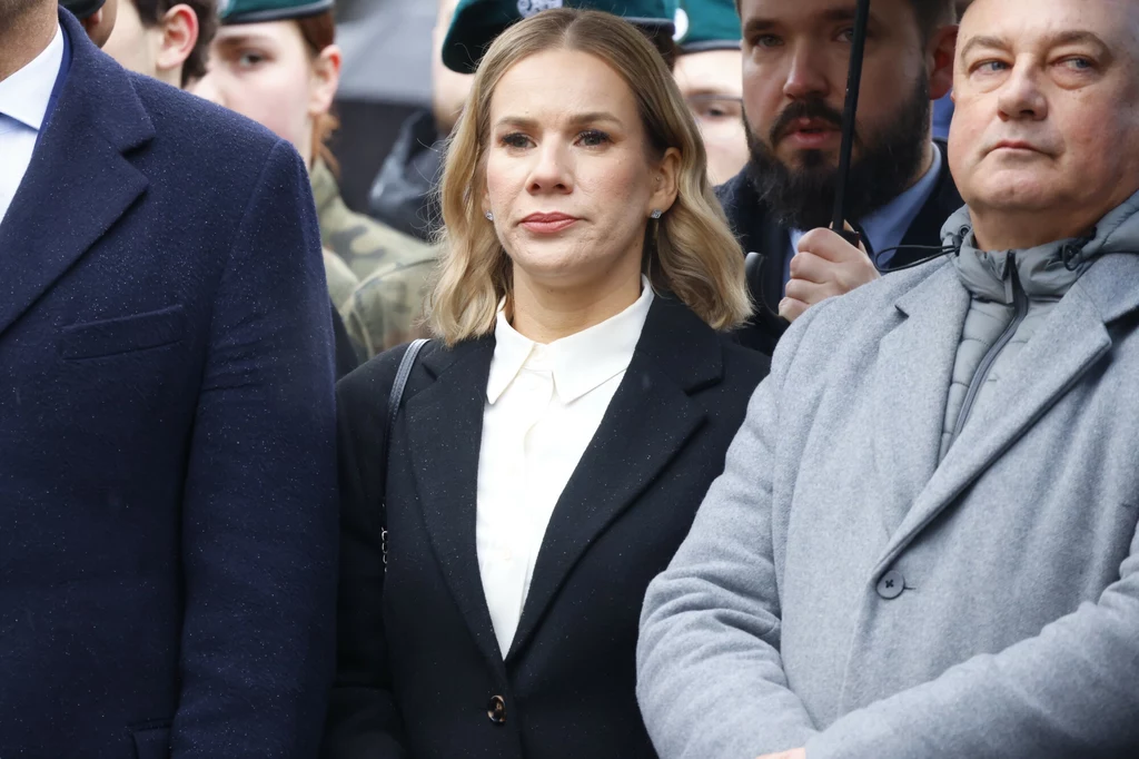 Narodowy Dzień Pamięci Żołnierzy Wyklętych z udziałem Marty Nawrockiej Kobieta w ciemnym płaszczu i białej koszuli stoi wśród innych osób ubranych elegancko, w tle mundurowi oraz tłum, atmosfera wskazuje na uczestnictwo w formalnym wydarzeniu.