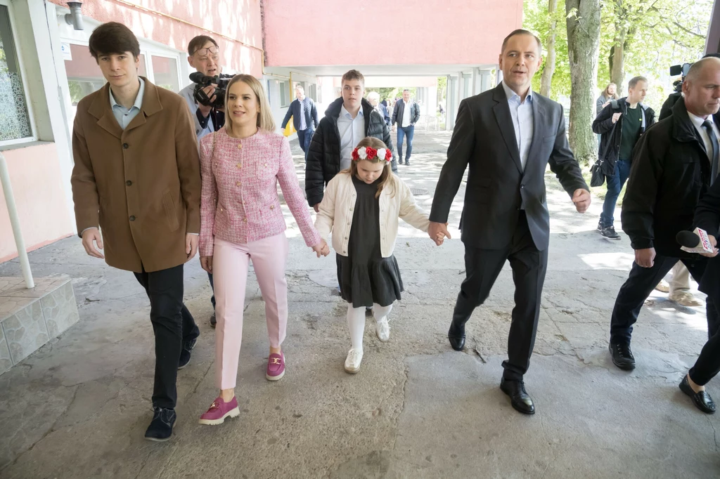 Marta i Karol Nawroccy oddają głos w I turze wyborów Grupa osób, w tym mężczyzna w garniturze, kobieta w różowym swetrze oraz dziewczynka z wiankiem we włosach, spacerują razem chodnikiem w otoczeniu innych ludzi, na tle budynku z różową elewacją.
