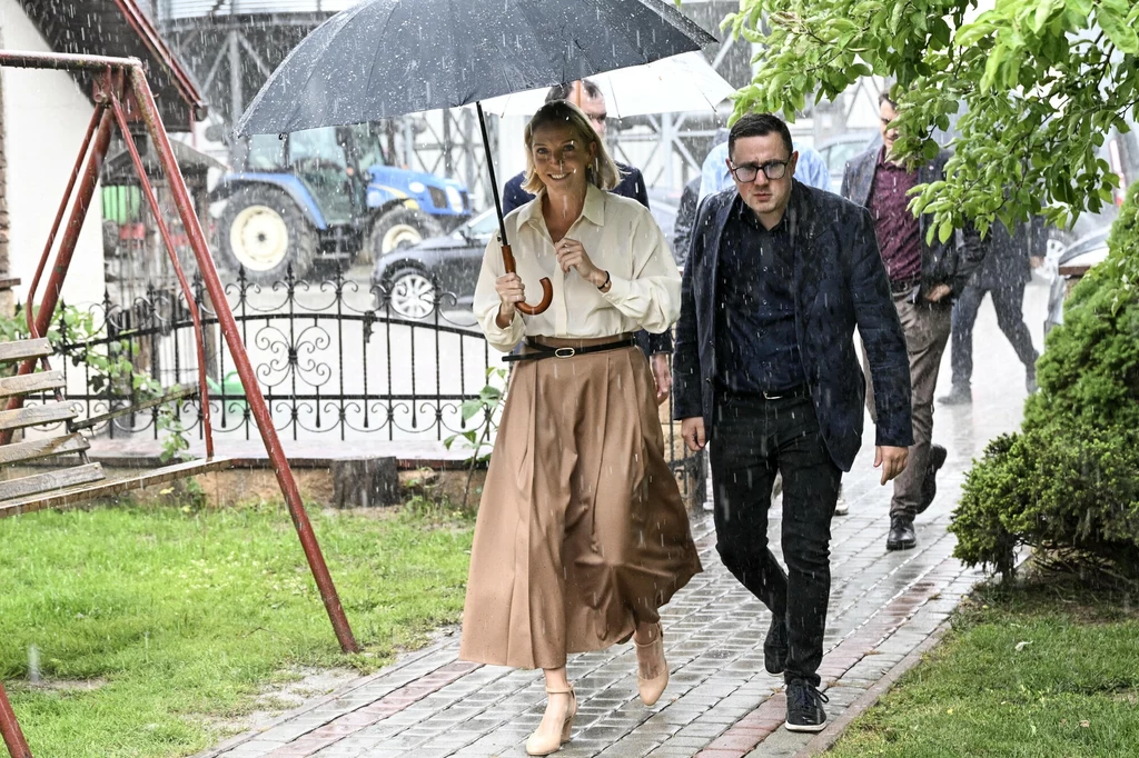 Małgorzata Trzaskowska w Niecieczy Dwie osoby idą razem chodnikiem podczas deszczu pod dużym czarnym parasolem, w tle widoczne są maszyny rolnicze i fragment placu zabaw.