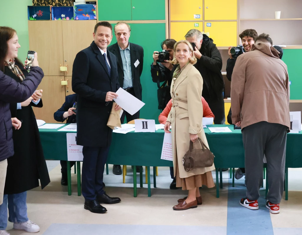 Marta i Rafał Trzaskowscy oddają głos w I turze wyborów prezydenckich Grupa osób przy stole komisji wyborczej, dwie osoby elegancko ubrane oddają głos, za nimi kilku fotoreporterów robi zdjęcia, jedna osoba korzysta z telefonu, wnętrze jest jasne z kolorowymi szafkami w tle.