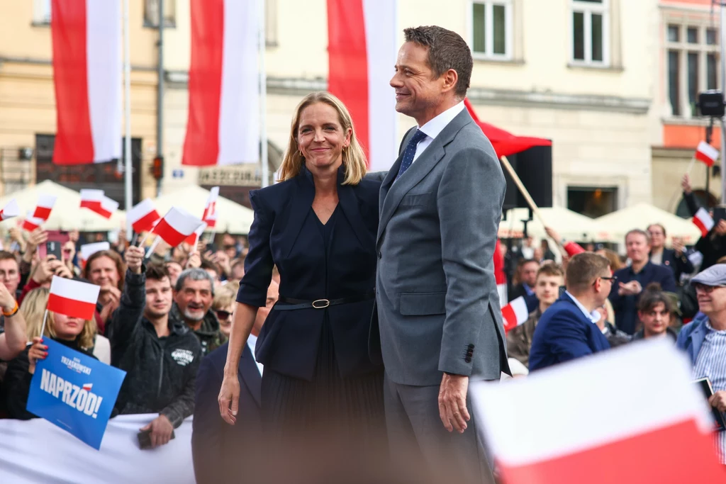Wiec Rafała Trzaskowskiego w Krakowie Tłum ludzi zgromadzonych na wiecu, dwie centralne postacie uśmiechają się do publiczności, wokół liczne biało-czerwone flagi i transparenty, w tle budynki miejskie.