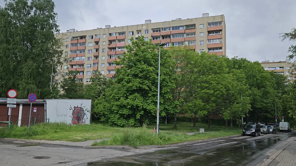 Prokocim, to tutaj mieszkali Jerzy i Jacek Wysoki blok mieszkalny z wielkiej płyty otoczony zielenią, z przodu widoczna niewielka altana, na której znajduje się graffiti, po prawej stronie zaparkowane samochody oraz asfaltowa droga z mokrym fragmentem po opadach deszczu.