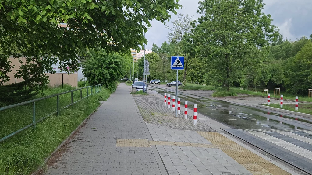 Prokocim, to tutaj mieszkali bohaterowie naszej opowieści Chodnik biegnący wzdłuż zieleni, barierka po lewej stronie, przejście dla pieszych z białymi pasami i słupkami ograniczającymi po obu stronach, znak przejścia dla pieszych oraz zaparkowane samochody w tle, otoczenie zielonych drzew i niskich budynków.