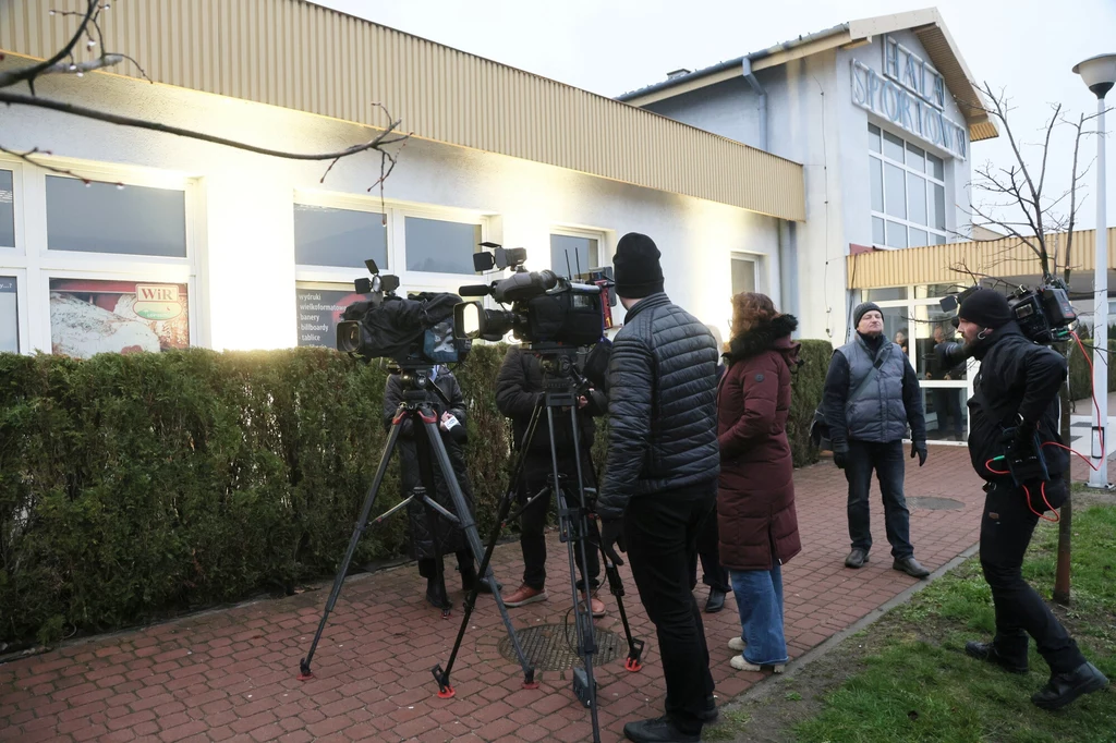Końskie, przedstawiciele mediów przed miejscową halą sportową, gdzie w kwietniu odbyła się debata prezydencka Grupa osób z kamerami i sprzętem nagraniowym stoi na chodniku przed niskim budynkiem o nowoczesnej fasadzie i dużych oknach, część uczestników ma na sobie ciepłe kurtki i czapki, wokół widoczne są zimozielone krzewy.