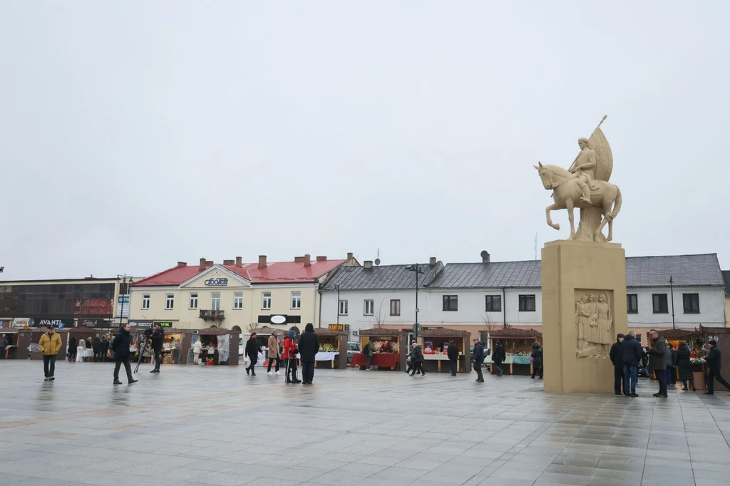 Rynek w Końskich Rynek miejski z niewielką liczbą osób spacerujących lub przebywających przed rzędem niskich budynków i stoisk targowych, po prawej stronie widoczny monumentalny pomnik przedstawiający postać na koniu.