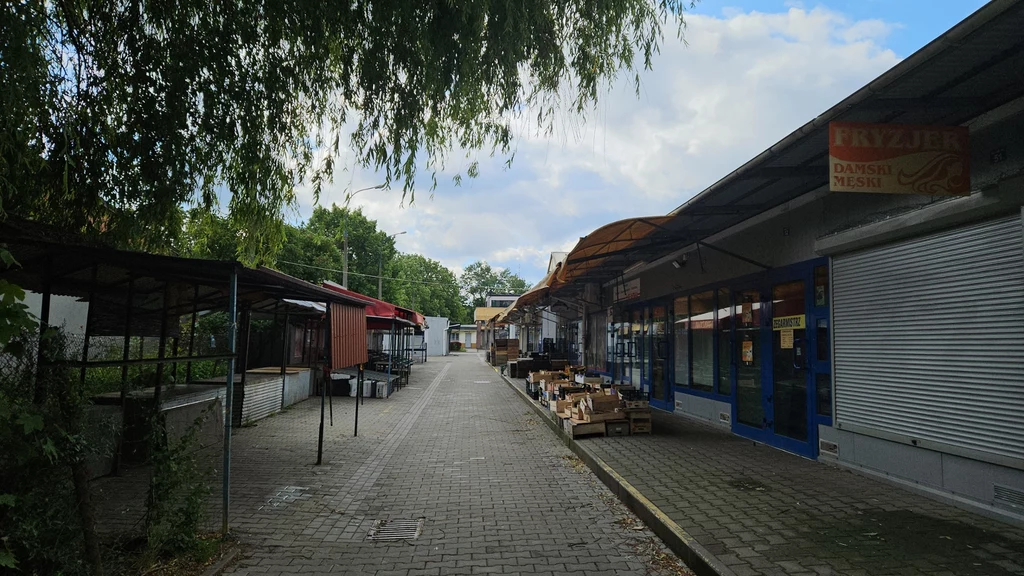 Plac, na który miał udać się Jacek, po telefonie od napastników Opustoszałe alejki targowe z zamkniętymi lub opuszczonymi stoiskami, po lewej stronie widoczne metalowe zadaszenia, po prawej zamknięte rolety sklepowe i puste kartony na chodniku, drzewa w tle dodają naturalnego otoczenia.