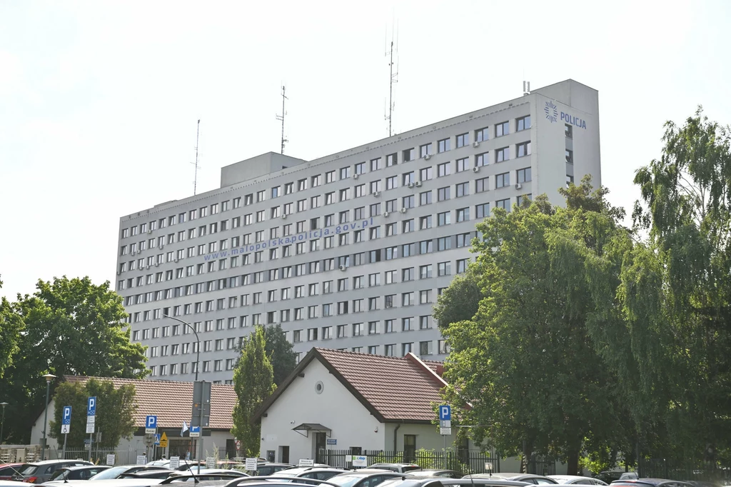 Komenda Wojewódzka Policji w Krakowie Wielopiętrowy budynek komendy policji otoczony drzewami, przed nim znajduje się parking z licznymi zaparkowanymi samochodami oraz niższy biały budynek w stylu stróżówki