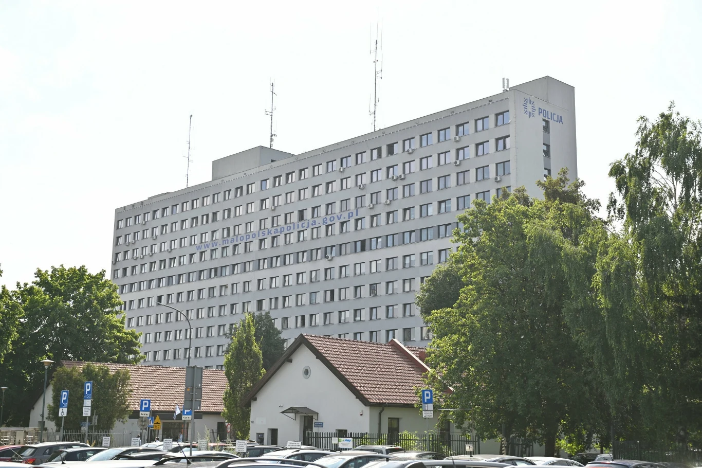 Komenda Wojewódzka Policji w Krakowie Wielopiętrowy budynek komendy policji otoczony drzewami, przed nim znajduje się parking z licznymi zaparkowanymi samochodami oraz niższy biały budynek w stylu stróżówki