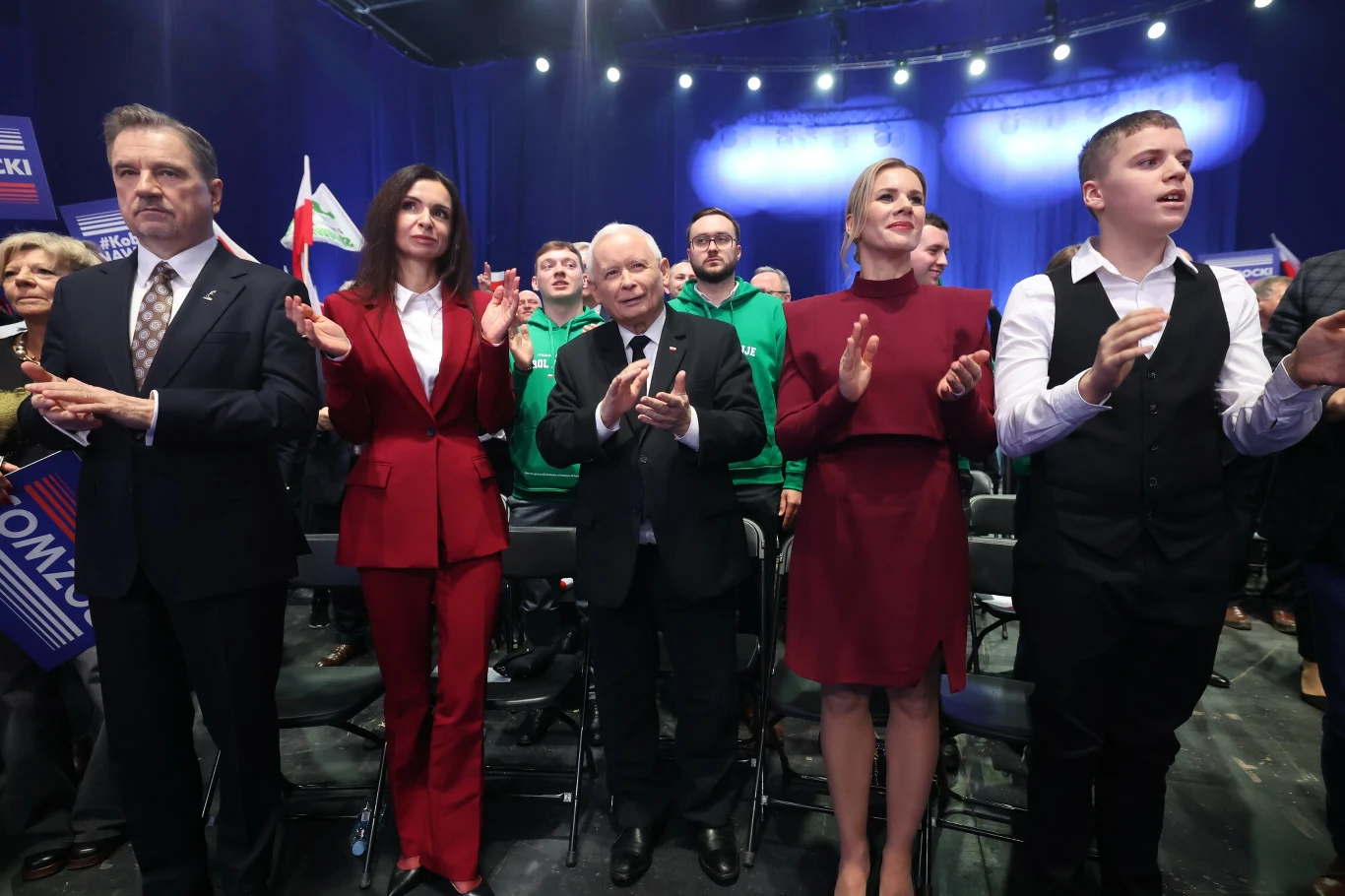 Grupa osób stojących w pierwszym rzędzie podczas wydarzenia politycznego, w tle widoczni uczestnicy oraz transparenty i flagi, oświetlone reflektorami.