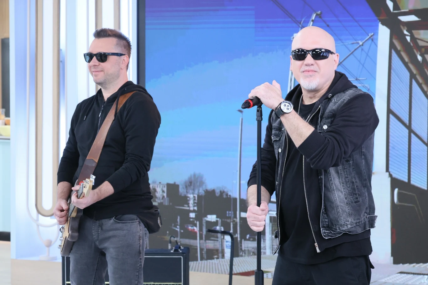 Dwóch mężczyzn w ciemnych okularach, jeden z gitarą elektryczną, drugi trzyma mikrofon stojąc przy statywie, obaj ubrani na czarno, w tle ekran z grafiką przedstawiającą fragment panoramy miasta i przewody trakcyjne.