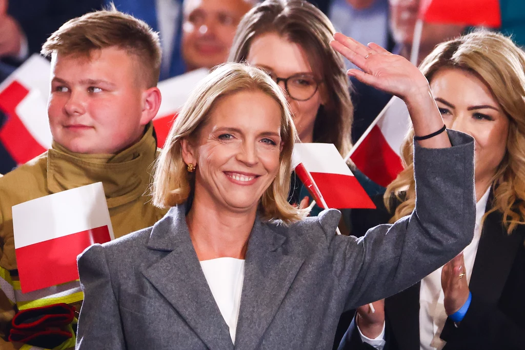 Małgorzata Trzaskowska jest absolwentką Uniwersytetu Ekonomicznego w Krakowie. Uśmiechnięta kobieta w szarym płaszczu macha ręką, wokół niej tłum osób z małymi flagami Polski, w tym młody mężczyzna w mundurze strażackim.