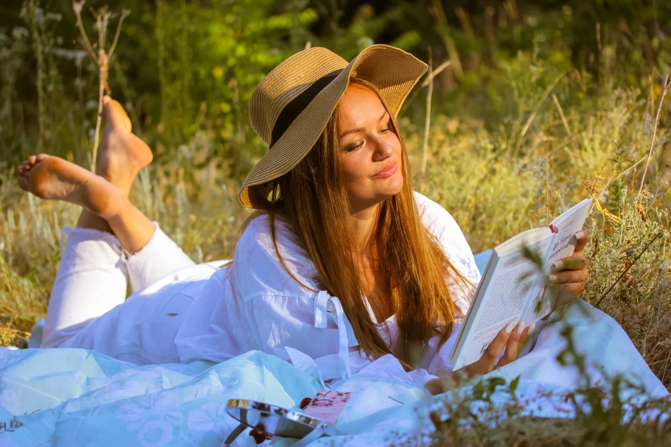 Człowiek-bluszcz. Uważaj na tę astrologiczną cyfrę, bo uzależni cię od siebie a potem porzuci Młoda kobieta w jasnym ubraniu leży na kocu na trawie w otoczeniu zieleni, czytając książkę i uśmiechając się, na głowie ma słomkowy kapelusz, wokół panować spokojna, letnia atmosfera.