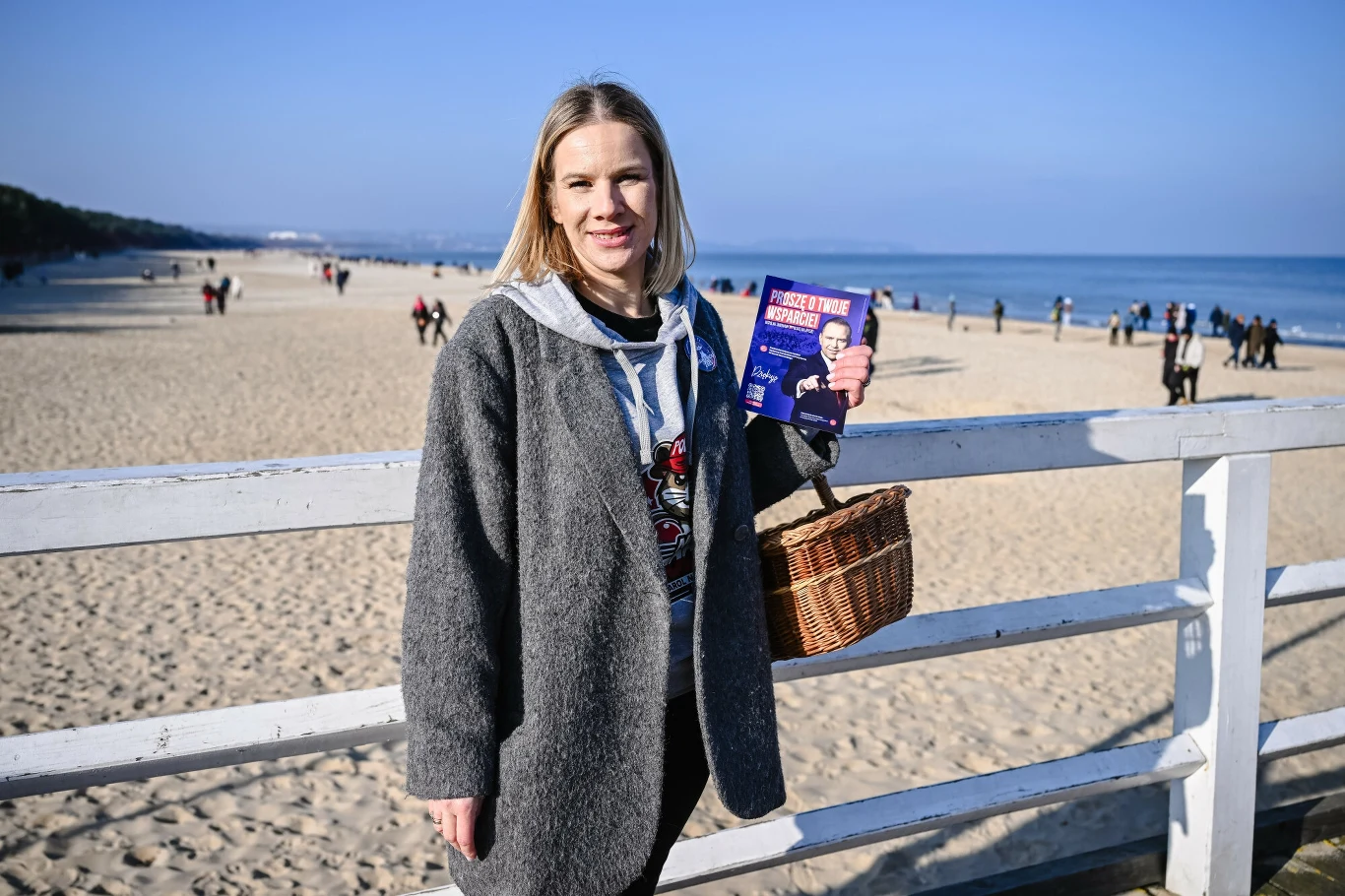 Kobieta w szarym płaszczu stoi na molo przy plaży, w jednej dłoni trzyma wiklinowy koszyk, a w drugiej ulotkę wyborczą. W tle widać ludzi spacerujących po plaży oraz morze. Marta Nawrocka