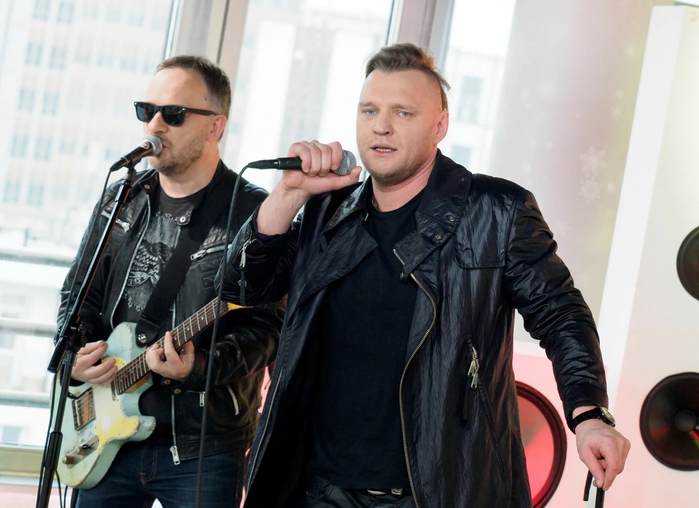 Dwóch mężczyzn ubranych na czarno występuje na scenie, jeden trzyma mikrofon i śpiewa, drugi gra na gitarze elektrycznej i śpiewa do mikrofonu, w tle widoczne duże okna i fragmenty sprzętu muzycznego.