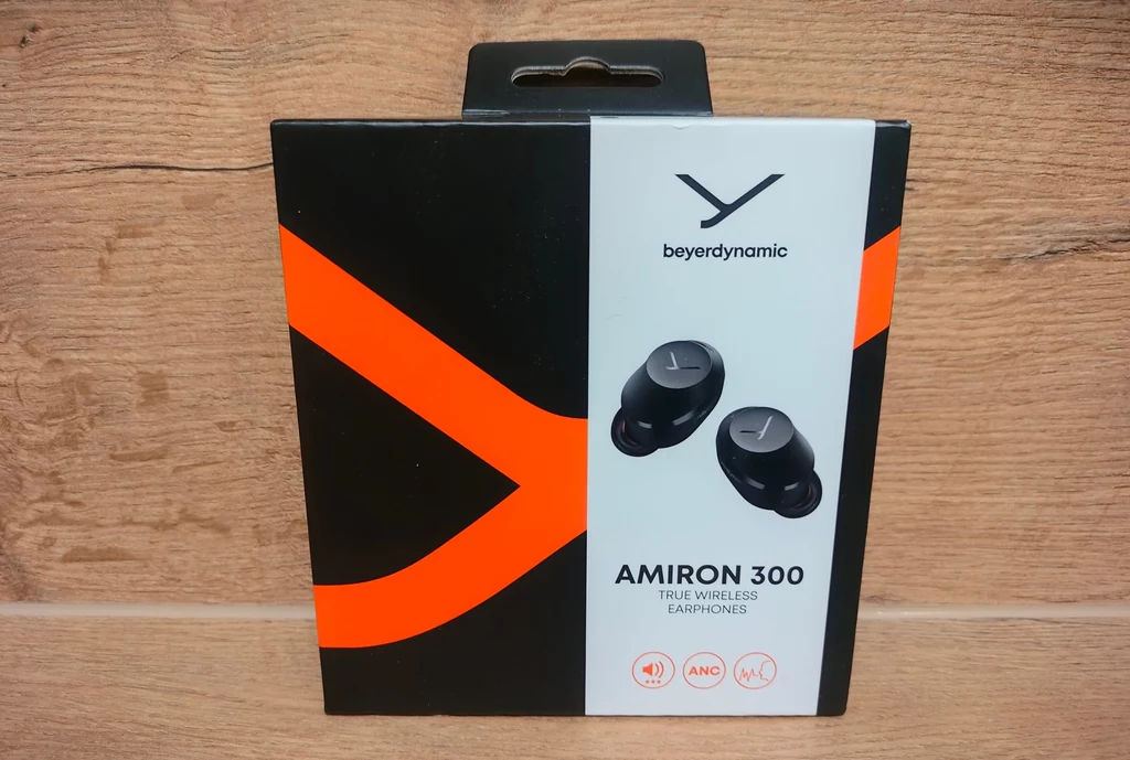 Beyerdynamic Amiron 300 Opakowanie bezprzewodowych słuchawek dokanałowych Beyerdynamic Amiron 300 na tle drewnianej powierzchni, dominują kolory czarny i pomarańczowy oraz widoczne logo producenta.