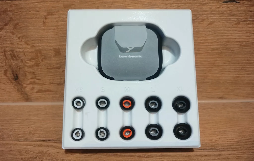 Beyerdynamic Amiron 300 Otwarty zestaw wkładek dousznych firmy Beyerdynamic z różnymi rozmiarami końcówek silikonowych w plastikowej przegrodzie oraz etui ochronne na drewnianym tle.